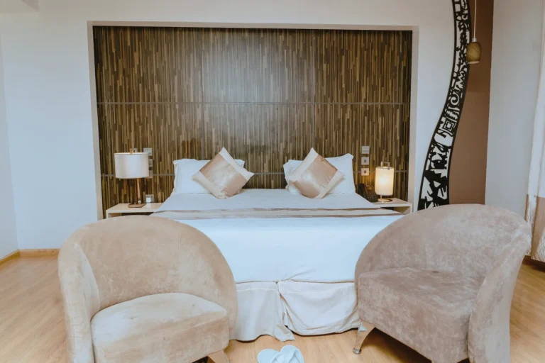 Deluxe room seen from the foot of the bed, with two velvet chairs, a timber headboard wall and a carved black screen