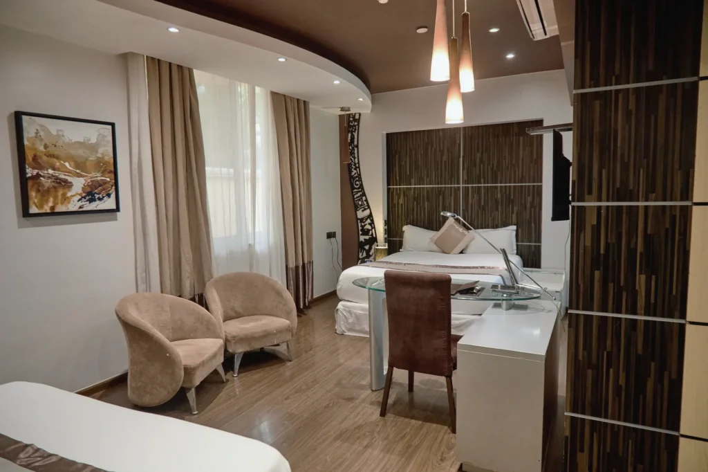 Twin Queen Room seen from the window side, with two velvet tub chairs, a glass writing desk and a timber headboard wall